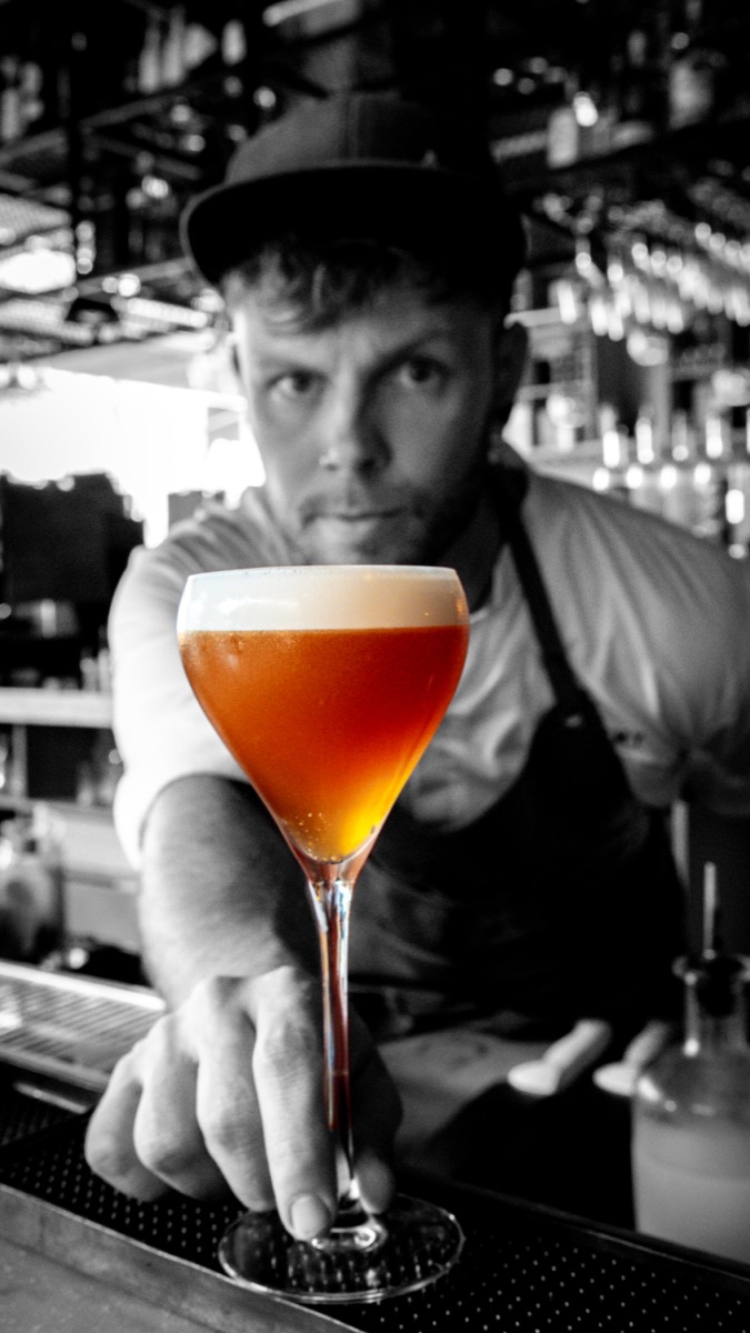 A striking monochrome image of a bartender presenting a vibrant orange cocktail, representing iconic digital creation and 100% original content.