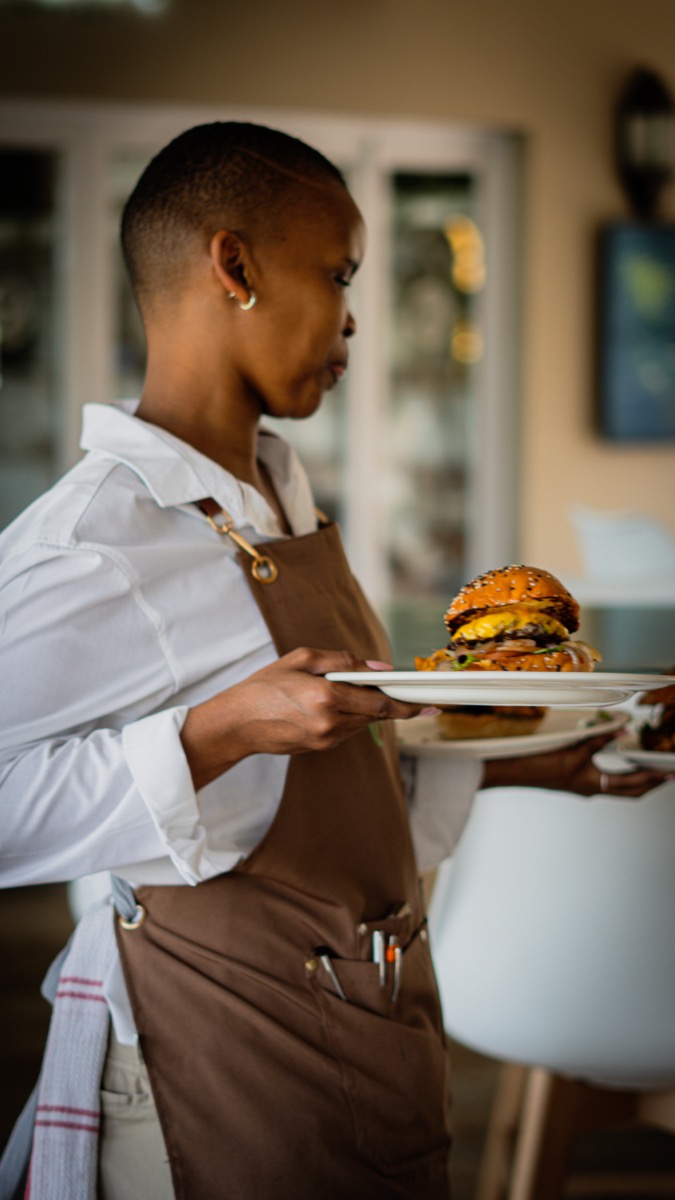 A focused server carefully carrying a plate with a large burger and other dishes, representing a content engine and scaling production for high-volume delivery.