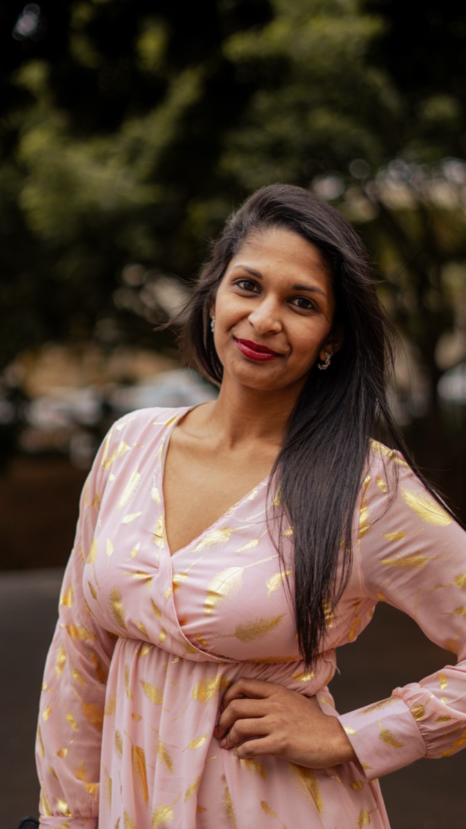 A smiling woman in a light pink dress adorned with shimmering gold feather patterns, representing 100% original work and a commitment to banning stock photography.