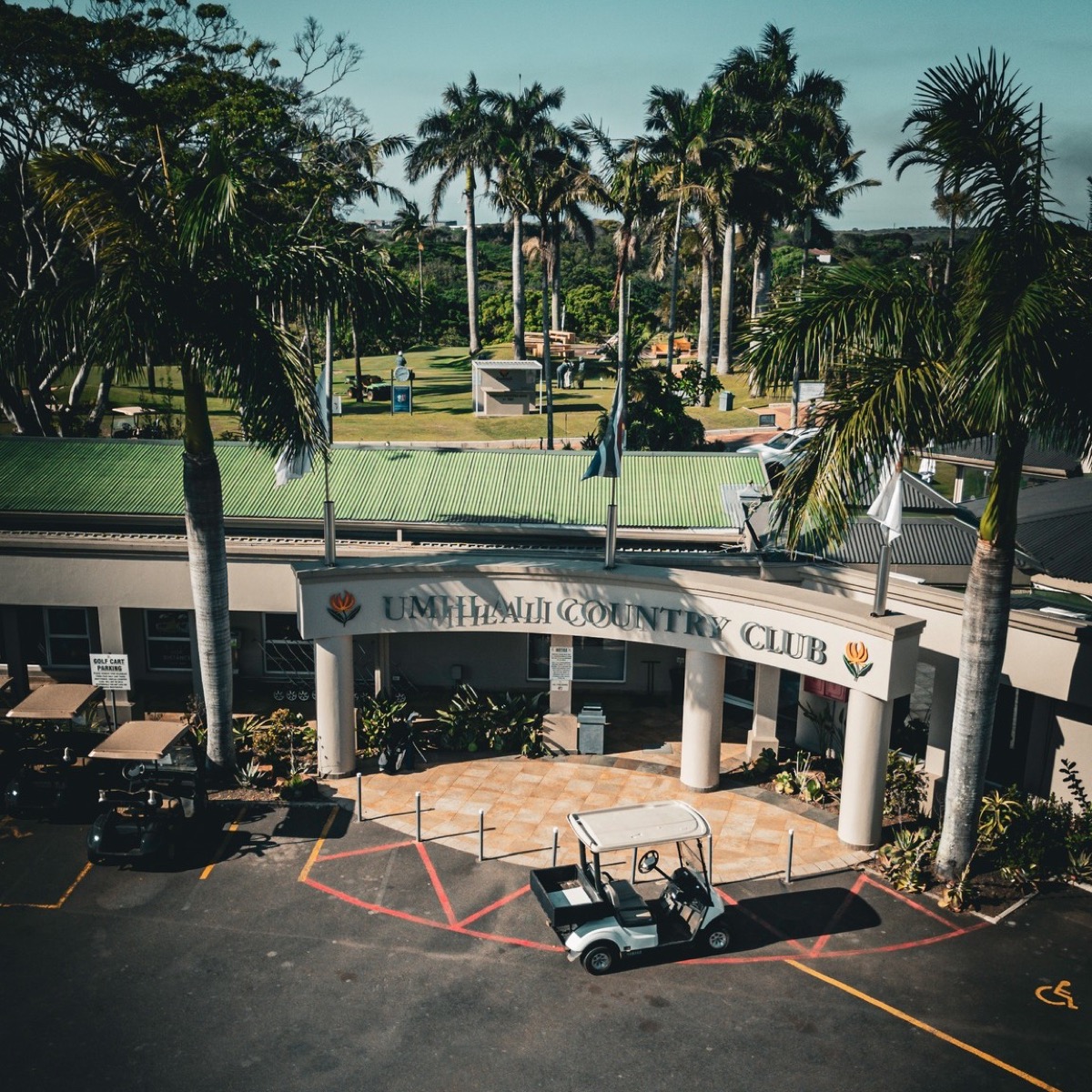 An aerial view captures the entrance of the Umhlali Country Club, featuring its arched sign, a white golf cart parked in the foreground, and numerous tall palm trees under a bright sky.